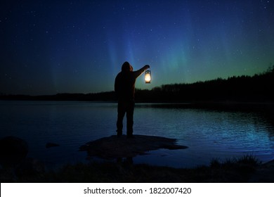 Silhouette of a man with a lantern near the lake. Night sky with stars and aurora borealis. Night landscape with northern lights and people.
