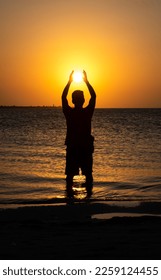 Silhouette of a man on the beach during sunset simulating catching the sun with his hands - Shutterstock ID 2259124455
