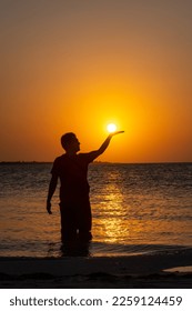 Silhouette of a man on the beach during sunset simulating holding the sun with one hand - Shutterstock ID 2259124459