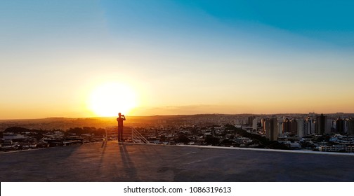 Silhouette Of A Man On Top Of A Building. Sunset On The City In The Background.