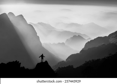 Silhouette Of Man On Top Of Mountain With Sunset In Black And White. Conceptual Scene.