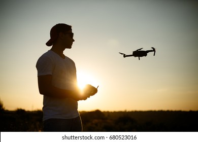 Silhouette Of A Man Piloting Drone In The Air With A Remote Controller In His Hands On Sunset. Pilot Takes Aerial Photos And Videos With Quad Copter From Above. Copter On Background