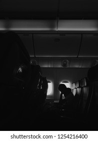 silhouette of man sitting against small window of airplane with book on his lap. Almost no light in the cabin. Guy has travel pillow on his neck.He propped up his head