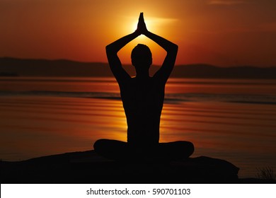 Silhouette Of Man Sitting In Lotus Pose, Holding His Arms Above Head In Namaste And Meeting Sunset