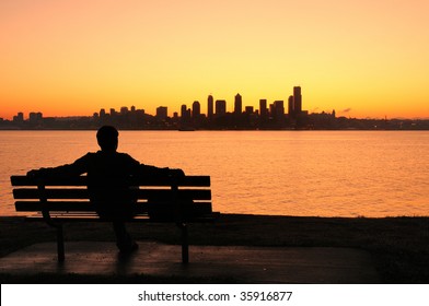 Silhouette Of A Man Sitting On A Park Bench Watching The Sun Rise Behind Seattle Skyline