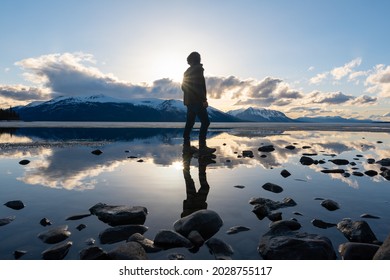 Silhouette of a man standing on a rocky lakeshore in northern Canada with reflection in calm lake below, dramatic clouds and blue sky. 