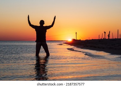 Silhouette Of Man At Sunset In Sea Water With Hands Raised Towards Setting Sun. Worship, Praise, Thanksgiving, Freedom And Love To Nature Concept