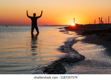 Silhouette Of Man At Sunset In Sea Water With Hands Raised Towards Setting Sun. Worship, Praise, Thanksgiving, Freedom And Love To Nature Concept