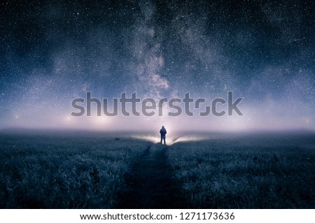 A silhouette of a man with a torch in a field, looking at lights on the horizon on a moody misty night. With a galaxy of stars overhead.
