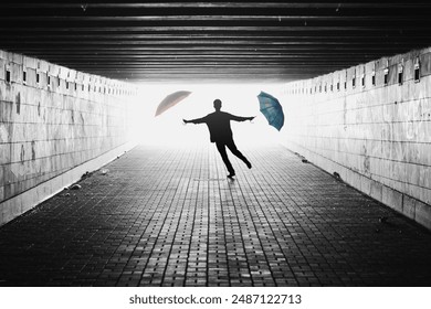 Silhouette of man with two umbrellas in a tunnel. Black and white image with light coming from behind. Person jumping towards the camera. Lens facing against the light. - Powered by Shutterstock