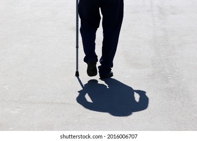 Silhouette Of Man Walking With A Cane Down The Street, Shadow On Asphalt. Concept Of Old Age, Diseases Of The Spine Or Joint Disease, Elderly People