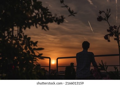 Silhouette Of Man Watching Sunset On Urban Rooftop Garden. Male Standing On Apartment Balcony Terrace And Looking At City At Sunset. Person Enjoys Evening On Skyscraper Green Roof During Dawn.