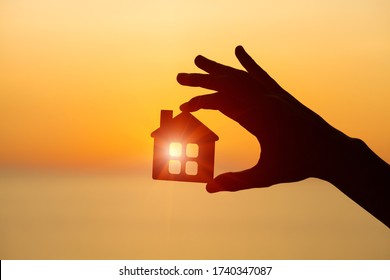 Silhouette Of Man's Hand Holding Small Wooden House Against Sunset Or Sunrise Light, Sweet Home And Family Concept, Solar Energy, Real Estate Agent Offer New House For Rent Or To Buy, Copy Space 