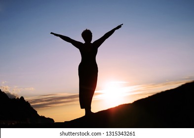 Silhouette of a mature woman with her arms spread out to the side, like wings, against the sunset sky