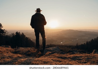 A Silhouette of a Middle Eastern man in a hat standing on a hilltop watching the sunset over a scenic valley in hiking area Bavarian Forest in Germany