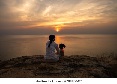 Silhouette mother and daughter sitting against sunset at the cliff of the sea.