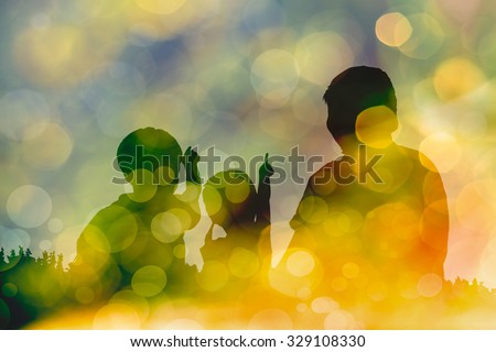 Silhouette of mother and two kids. Little children point to sky. Twinkling lights vivid blurred bokeh abstract in yellow background. Friendly family.