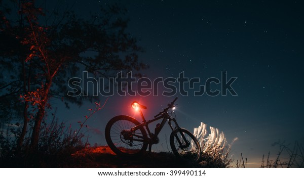 Silhouette of mountain bicycle\
on a background of the sky. \
Bicycle stands on the hill, while\
cyclist rests.\
Headlamp and taillight are glowing on the mountain\
bike.\
\
