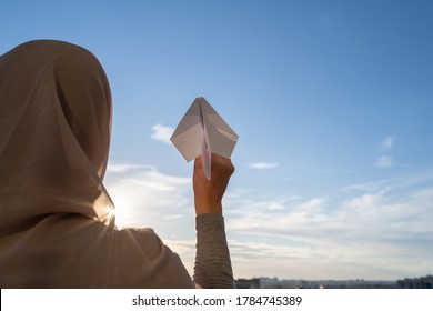 Silhouette Of Muslim Woman In Head Scarf Launching A Paper Airplane On Blue Sunset Sky Background. Travel Concept 