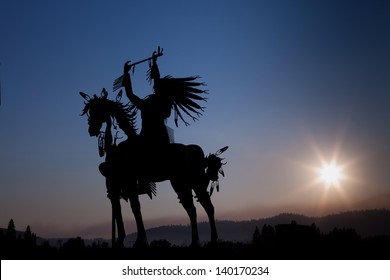 A silhouette of a Native American on a horse made from metal with eight rays emanating out from the setting sun in the distance above hazy mountains.