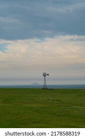 A silhouette of an old fashioned windmill stands alone along the horizon of a prairie with a butte in the background during a cloudy rainy day in North Dakota. 