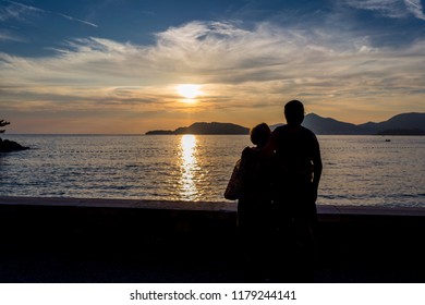 Silhouette Of Older Couple Watching Sunset On Vacation. Sun Going Into Sea With Beautiful Clouds