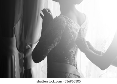 Silhouette of one young slim bride dressing with help of female hands in lace wedding dress preparing for ceremony standing near window black and white, horizontal picture