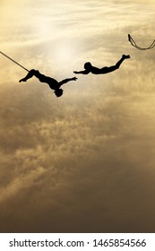 Silhouette of a pair of trapeze artists swinging through a surreal cloudy sky. 