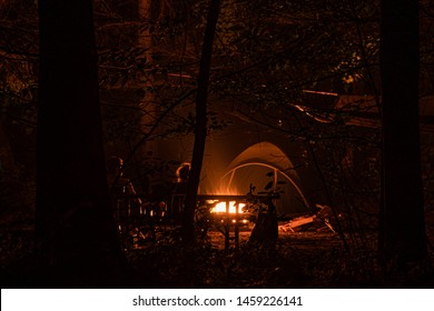 Silhouette Of People Sitting Next To Fire At A Camp Site With A Tent In The Background. Night Shot, Long Exposure. Hiking, Camping, Outdoor Lifestyle Concept.