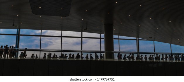 Silhouette Of People Standing In Line On Upper Floor Of Airport Building In Front Of Blue Sky Outside Window. Selective Underexposure For Effect.