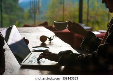 Silhouette Of Person Working On Computer Drinking Coffee At Wooden Table At Hotel Lobby With Forest On Background Throw Large Windows