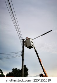 Silhouette Photo Electrician Working On Power Line. Technician On Crane Bucket Truck High Up Of A Crane To Fix Main Voltage Cable In Housing Electrical Pole While Rain Clouds Are Coming.