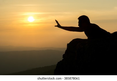 A Silhouette Photo Of A Man Laying Down By The Edge Of The Cliff Reaching Out His Arm Trying To  Grab The Sun 
