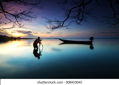 Silhouette of photographer looking to the boat near the beach when the sun goes down with branches tree border. Nature Photography