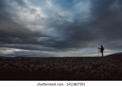 Silhouette of a photographer or traveler with tripod standing on stone. Background of a dramatic sky. Hazardous working conditions and human silhouette - Shutterstock ID 1411807391