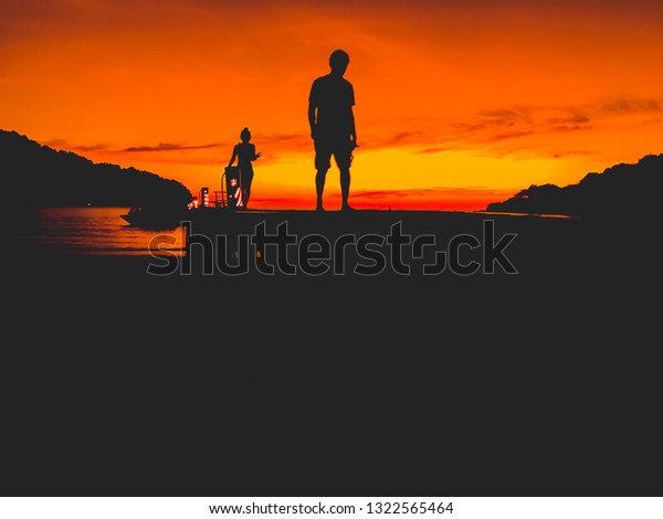 Silhouette picture of couple\
walking on the bridge during the sunset with orange sky in evening,\
Two peoples walking on the wooden jetty (pier) extending to the\
sea.