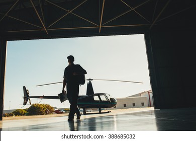 Silhouette Of A Pilot Arriving At The Airport With A Helicopter In Background. Helicopter Pilot In Airplane Hangar.