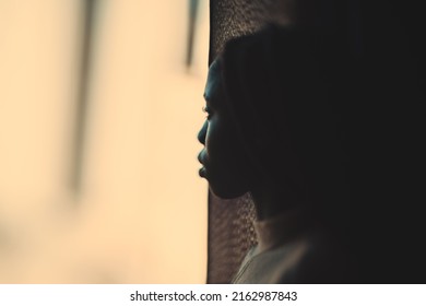 A silhouette portrait of a cute young black female pensively looking outside the window while standing next to the curtains, selective focus on her eyelashes, shallow depth of field