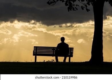 Silhouette portrait of a lonely anonymous man sitting on a bench watching the dark skies above roll by. 