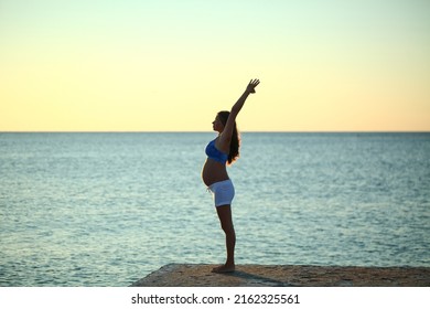 Silhouette pregnant girl doing yoga on the seashore in the morning against the sky and water. A woman does exercises on the beach background of sunset sunrise. Asanas from the sun greeting complex