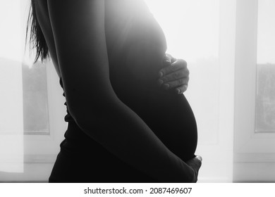 Silhouette of pregnant woman in dress at home by window touching belly. Black and white foto close up