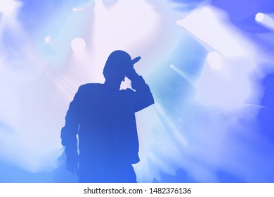 Стоковая фотография молодого рэп-певца с микрофоном в руке, поющего популярную песню на сцене в синих огоньках. Хип-хоп исполнитель выступает вживую на сцене в мюзик-холле.