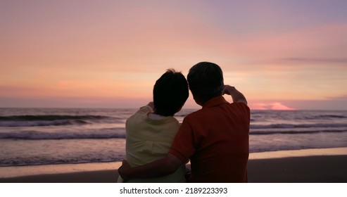 Silhouette Rear View Of Asian Elderly Couple Sitting On The Beach And Chatting Happily In Sunset Background