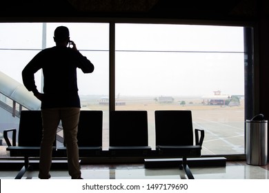 The Silhouette Of The Rear View Of A Man Making A Phone Call While Standing In Front Of A Glass In An Airport Waiting Room