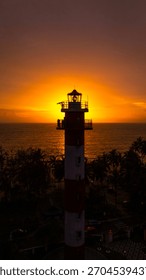 Silhouette of a red-white lighthouse during a dramatic sunset by the Arabian Sea, with golden light glowing behind the tower and palm trees along the coastline.