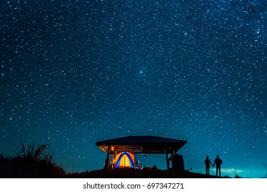 Silhouette Of Romantic Couple Watching Sky Full Of Stars On Mountain.  Glowing Tent And Lovers With A Starry Night View.