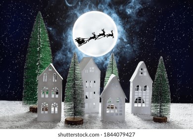 Silhouette Of Santa Claus With Bag Of Presents In A Sleigh With Reindeer Over Moon Above The Snowy Paper Village. Christmas Greeting Card.