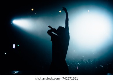 Silhouette of singer rising his hand opposite stage crowd. Floodlights lightning at the camera. Beautiful pool of white light on the background. - Powered by Shutterstock