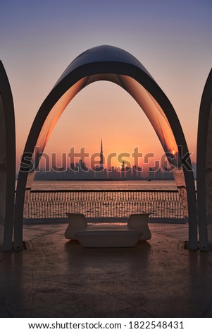 A silhouette of skyline of Dubai at sunset framed by an arch-shaped seating shade.                             