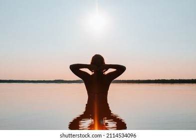 Silhouette Of Slim Young Woman Swimming In Lake At Sunset, Standing On Water And Enjoying Outdoor Recreation In Nature, Back View